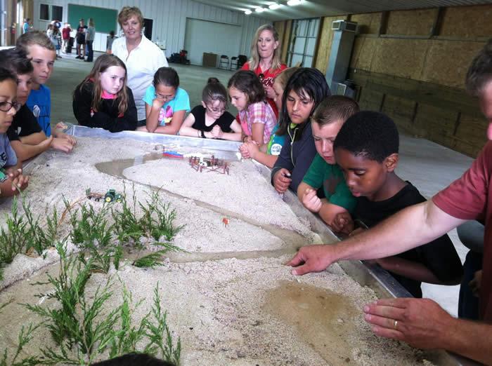 Kids stand around stream table while instructor shows them how water causes erosion.